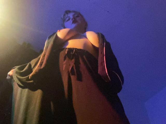 CocoVolkov, low-angle shot, wearing a loose robe, exposing her big, bare tits with pink nipples, brown pants, and dark hair, under blue and yellow lighting.