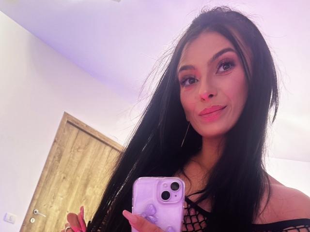 KinkyAmelie mit langen schwarzen Haaren, rosa Lidschatten und rosa Lippenstift, lächelt in die Kamera, hält ein rosa Telefon, trägt ein schwarzes Netzoberteil und zeigt ihre Brüste..