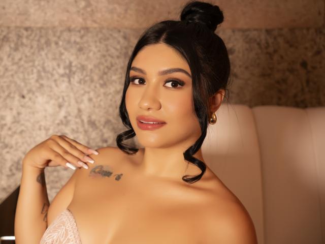 CamilaVerone has dark hair in a bun, brown eyes, and a tattoo on her right shoulder. She's wearing a lace strap, looking at the camera with a slight smile.