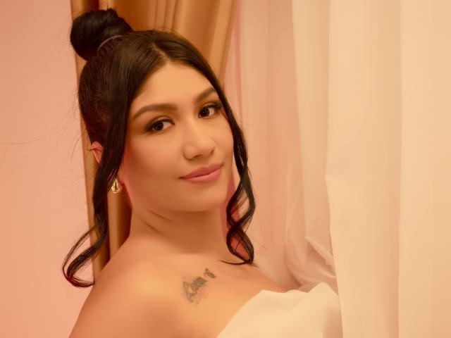 CamilaVerone with dark hair in a bun, wearing a white strapless top, has a tattoo on her chest, small earrings, and a subtle smile, against a pink and white backdrop.
