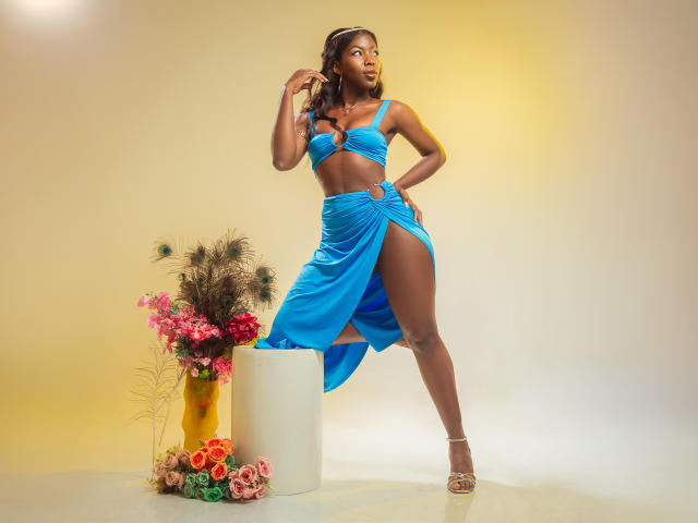 MichelleFlores, donkerhuidig, draagt een blauwe jurk met uitsparingen die haar middel blootlaat en een hoge spleet, staat op hakken, met haar hand in haar haar, en poseert naast een gele vaas met bloemen..