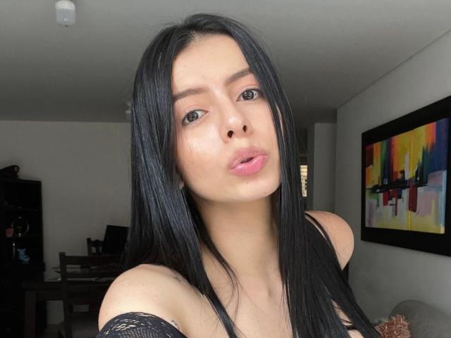 IsabellaJames has long black hair, pouty pink lips, and light skin, wearing a black lace top, looking at the camera with a slight pout. Her tits are visible.