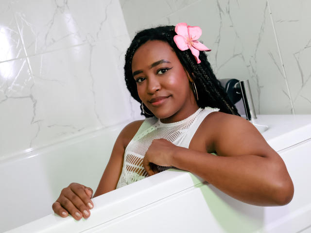 NaomiReyes with dark skin, braided hair, pink flower, white mesh top, in bathtub, smiling, holding tub edge, showing her tits, looking cute and relaxed.