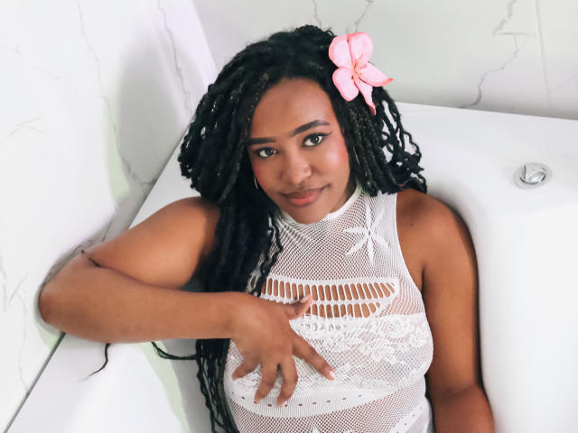 NaomiReyes with dark skin and long black braids, wearing a white mesh top, sitting in a bathtub, pink bow in hair, right hand on her large, lace-covered tits.