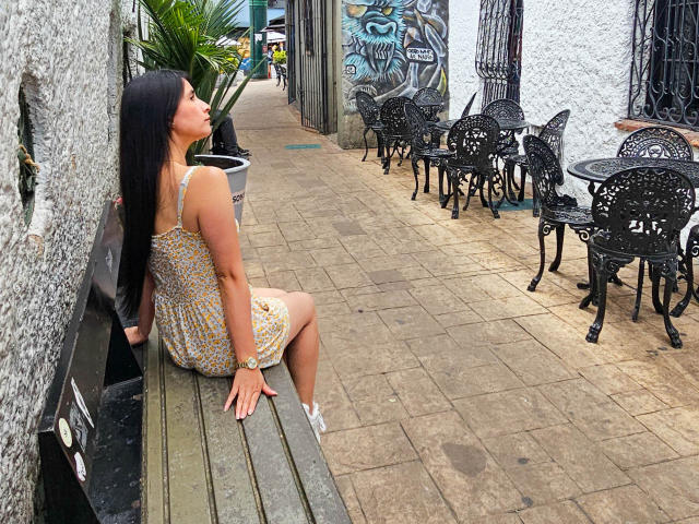 JainaRogers is sitting on a wooden bench in a floral dress, looking up, with black hair, medium tits, and white sneakers, next to black metal chairs on a stone sidewalk.