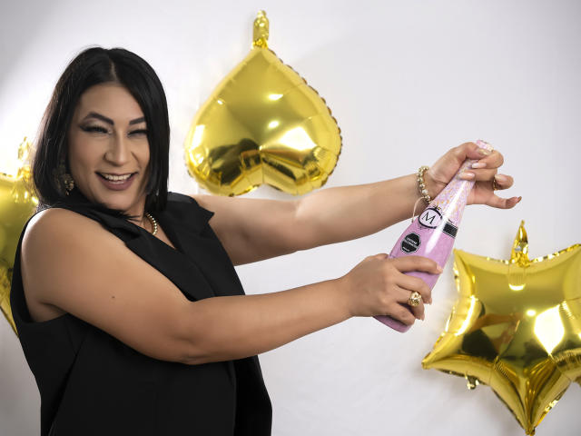EmaFoxy with black hair, smiling, holding a pink vibrator, wearing a black sleeveless top, gold star balloons in the background. She's smiling, showing her tits, and has a gold bracelet.