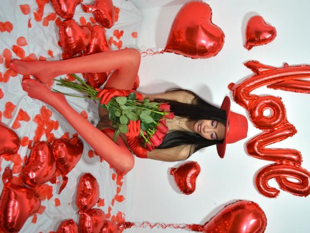 Adaallen with long black hair, wearing red hat and gloves, sitting with red heart balloons, holding roses, in red stockings, looking at camera, surrounded by red heart-shaped balloons.