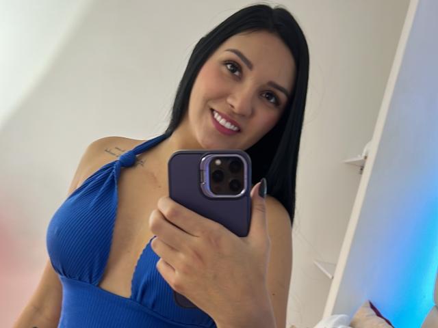 YessiJaymes with black hair, smiling, wearing a blue halter dress that reveals her large tits, holding a purple phone, visible tattoo on left shoulder.
