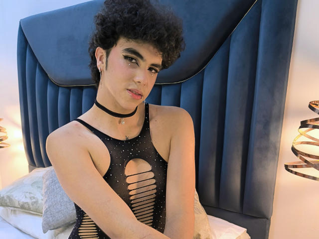 KheylerThompson, met krullend donker haar, draagt een zwarte top met gaas en uitsparingen, een zwarte choker en make-up, en zit op een bed met een blauwe hoofdbord. Kheyler heeft kleine borsten en roze lippen..