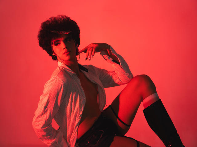 KheylerThompson avec des cheveux afro, chemise blanche ouverte, sans soutien-gorge, short noir, bas jusqu'aux genoux, éclairage rouge, main sur la joue, assise, regardant la caméra, confiante, sexy, poitrine dénudée..