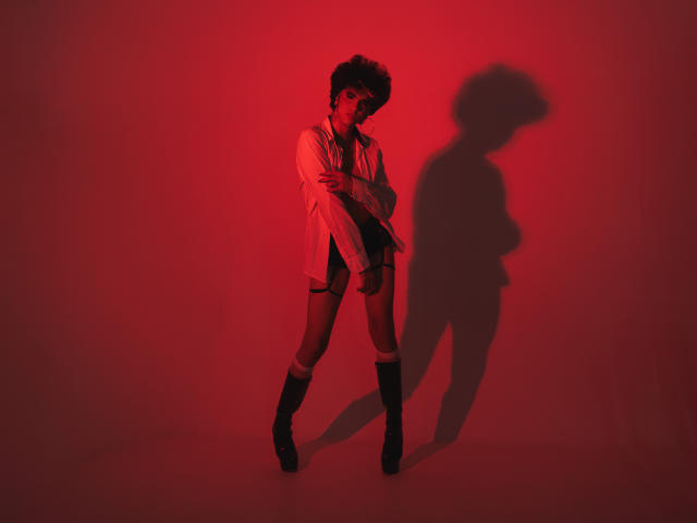 Kheyler Thompson, avec ses cheveux courts et foncés, portant une chemise blanche ample, un short noir, des chaussettes montantes et des bottes, projetant une ombre avec un éclairage rouge, debout, les mains près de sa poitrine..