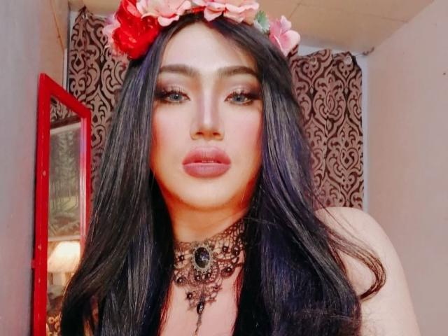 FancyShaine with long black hair, pink flower headband, and dark makeup, wearing a detailed necklace, has full lips and a soft pink blush, looking at the camera.