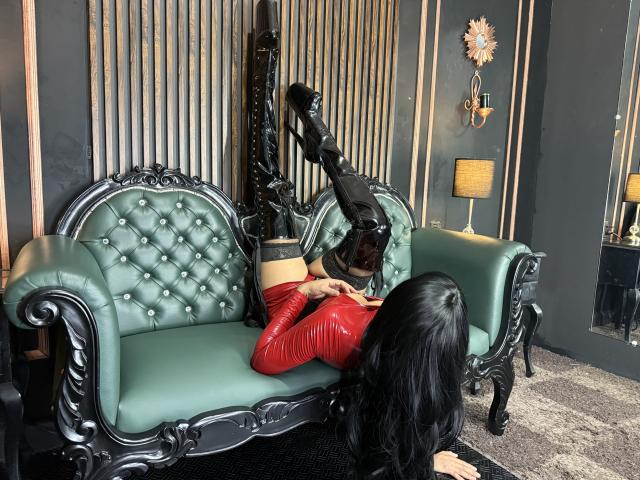 KarlaBella, with long black hair, is on her back on a green tufted sofa, wearing a red latex top and black boots, with her black boots in the air.