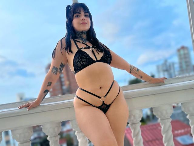 AmelieGracee, portant un soutien-gorge en dentelle noire et des culottes à fines bretelles, se tient sur un balcon, les bras reposant sur la rambarde, montrant ses tatouages, avec des cheveux noirs, un ciel bleu et des bâtiments de la ville en arrière-plan..