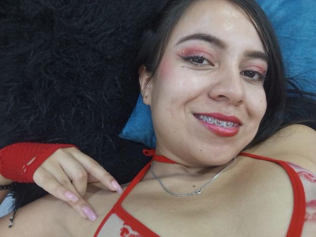 JessyLenon with red eyeliner, pink lipstick, and metal braces, smiling, wearing red strappy top, fingerless red glove, pink nails, lying on black and blue textured surfaces.