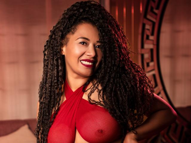 KenyaKarath with long, curly black hair, red mesh top revealing her large, round tits, and red lipstick, smiling at the camera. Warm, red lighting highlights her confident, sexy look.