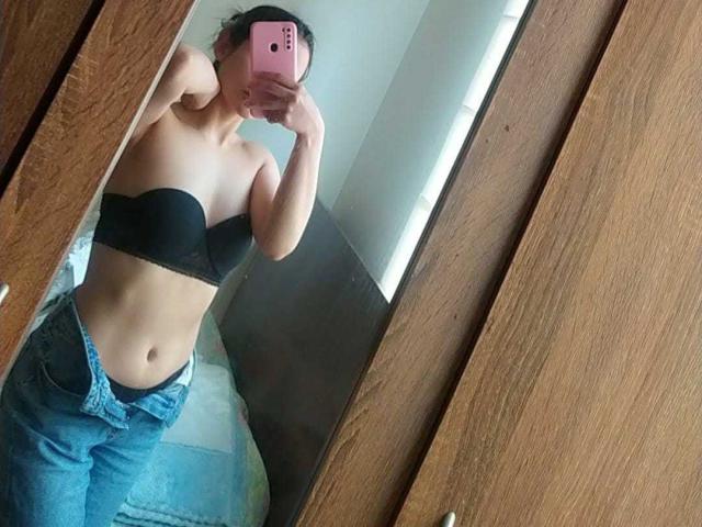 TinyLaurent taking a mirror selfie, wearing a black strapless bra and unbuttoned jeans, showing her flat stomach and small tits, holding a pink phone, in a wooden room.