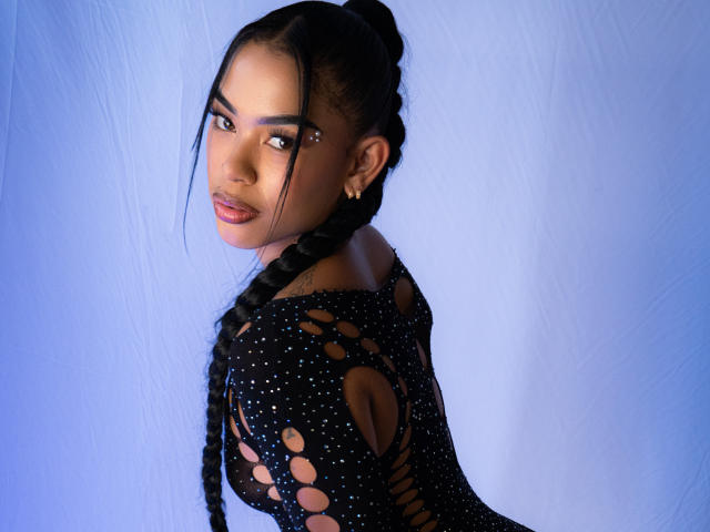 ScarletSummer, con cabello oscuro trenzado, vestida con un vestido negro, con cortes y lunares, que deja ver sus pechos a través de los agujeros, mirando por encima del hombro, iluminación azul y aretes dorados..