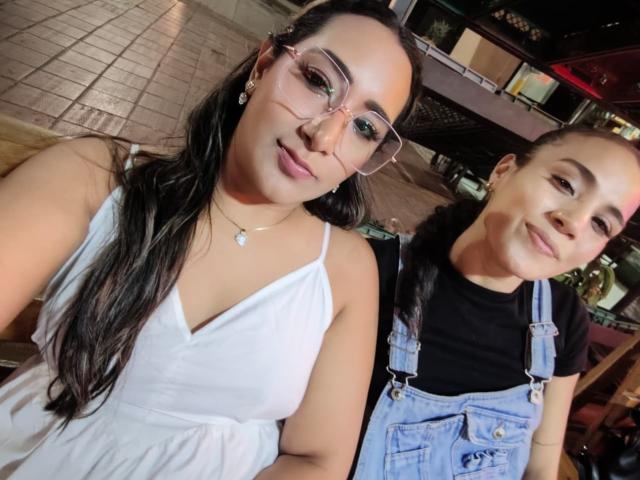 Isabela met een bril, een witte jurk en een gemiddelde borstmaat, staat naast Alahia in een denim overall, beiden glimlachen. Ze hebben beiden lang, donker haar. Isabela's neus is lichtelijk gekneusd. De setting is binnen..