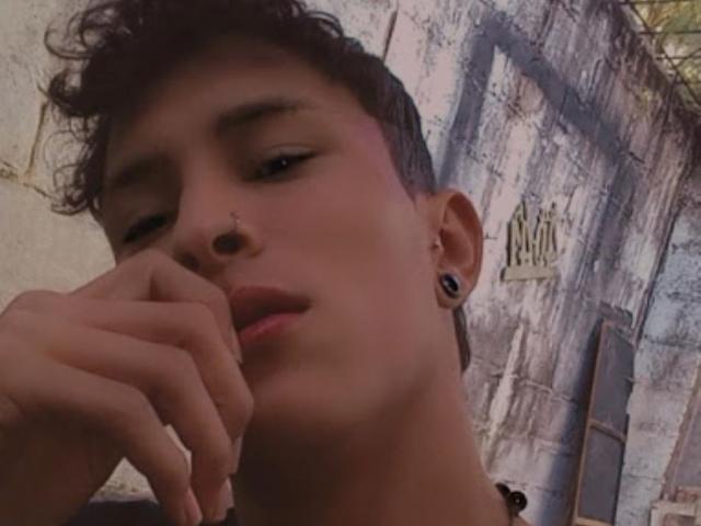 ErosAndree has short brown hair, a nose ring, black ear gauges, and is slightly touching their chin with a thoughtful expression in a dimly lit, gritty setting.
