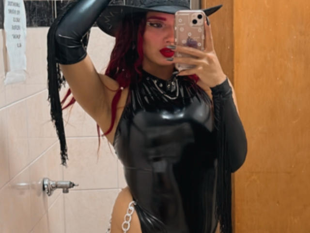 AlexaFoster in a black latex outfit, wearing a witch hat, long red hair, red lipstick, black gloves, holding a phone, showing her shiny, tight, latex-covered tits, and a chain belt.