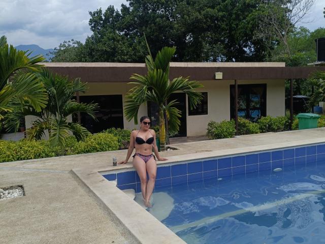 HanaMariana, com seios grandes, em um biquíni preto, em pé em uma piscina com azulejos azuis, usando óculos de sol, segurando uma bebida e olhando para a câmera. Ela também tem uma traseira atraente..