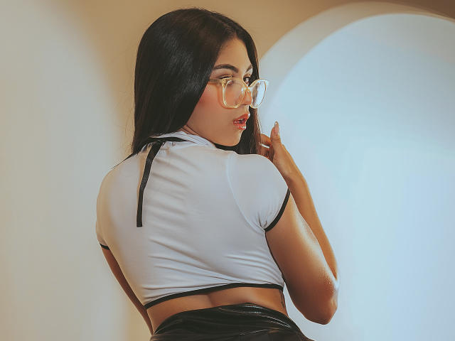 AnaWillsson with long black hair, wearing clear glasses and a tight white crop top showing her back, black latex skirt, and her hand near her mouth, looking over her shoulder.