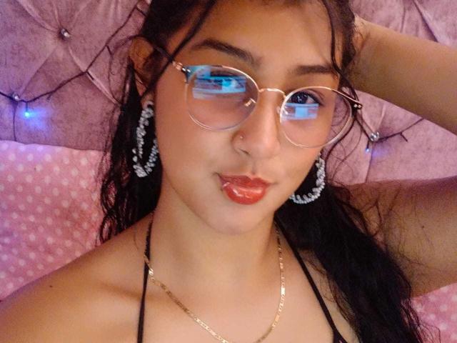 EmiilyJohamson with round glasses, puffy red lips, black hair, white pearl earrings, gold necklace, and black halter top, looking at the camera with a hand behind her head.