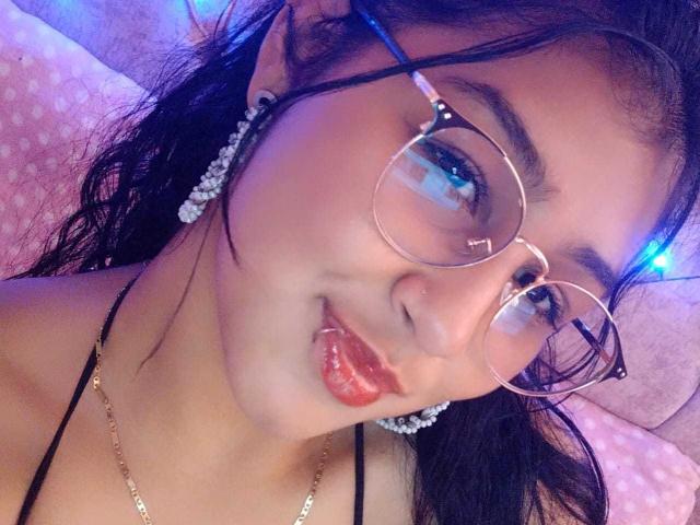 EmiilyJohamson with puffy red lips, wearing round glasses, black straps, and pearl earrings, sticking out tongue, blue light in background, wavy black hair, and gold necklace.