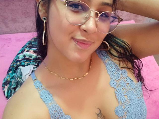 Emiily Johamson, con gafas, aros grandes, cadena de oro, blusa de encaje azul, piercing en la lengua y cabello oscuro y húmedo, sonriendo a la cámara, mostrando pequeños senos y un tatuaje en el pecho..
