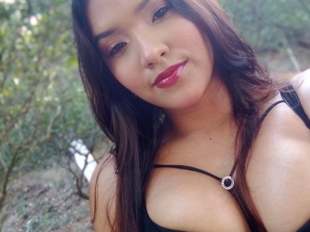MissLauren with medium tits, brown hair, and glossy pink lipstick, wearing a black top with a silver necklace, smiling in a forest. Her skin is tan, and she's cute.