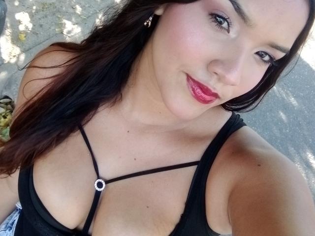 MissLauren with dark hair, red lipstick, and black strappy top, showing her tits and a small silver ring near her cleavage, smiling at the camera.