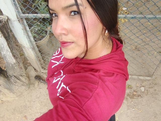 MissLauren with dark hair, pink lipstick, and red hoodie, showing her tits, standing near a chain-link fence, looking at the camera with a slight smile.