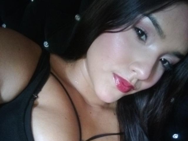 MissLauren's face with glossy pink lips, dark brown hair, and black tank top showing her large, shiny tits. She's looking at the camera with a slight, sexy smile.