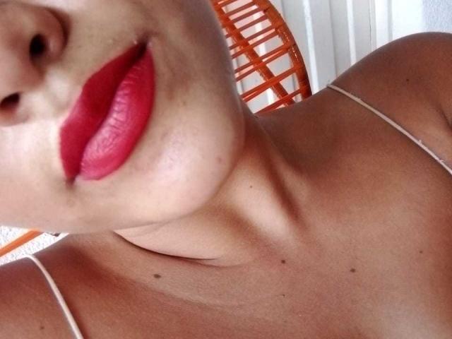 MissLauren's face with red lipstick, showing her plump lips and smooth, tan skin. Her collarbone and a thin strap are visible. Orange chair in the background.