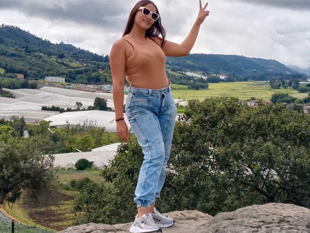 MissLauren with bare tits, wearing high-waisted jeans and white sneakers, standing on a rock, waving with her right hand, sporting sunglasses and long brown hair.