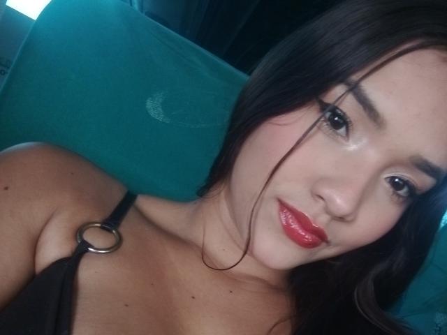 MissLauren with shiny red lips, black strap dress showing a ring, dark brown hair, and brown eyes, lying on a teal couch, showing her tits and small moles on her chest.