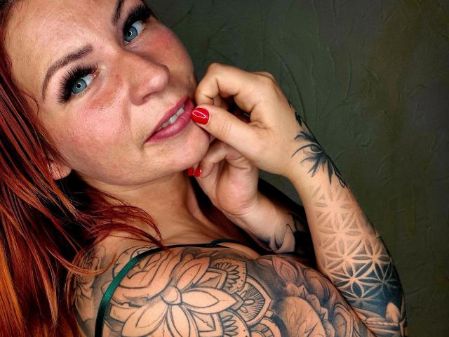 Chrissi69-hot with red hair, blue eyes, and red nails, biting her finger, showing tattoos on her arm, including a flower and geometric patterns. She has freckles and a cute smile.