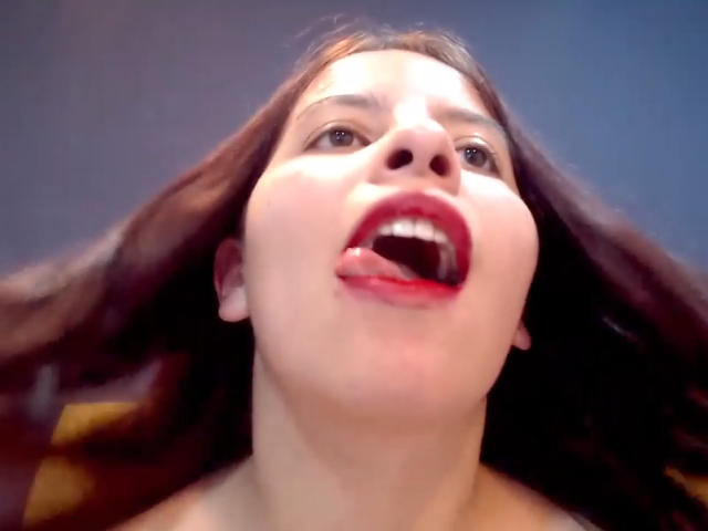 JuliaWaine is lying naked with her tits exposed, legs spread, and her hand on her pussy. She's sucking red lipstick and has a bracelet on her wrist.