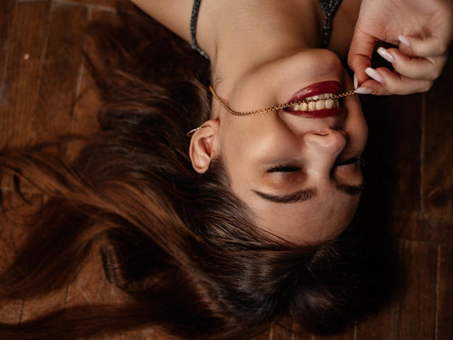 SalomettX with eyes closed, has large tits, and light brown, wet, shoulder-length hair, sitting topless on a bed, light skin, slight smile, soft background, casual, relaxed.