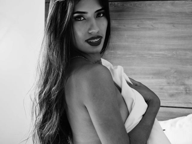 AnnyMayers with long dark hair, dark lipstick, and a seductive smile, holding a white sheet to cover her tits. Black-and-white photo, her eyes are half-open, looking at camera.