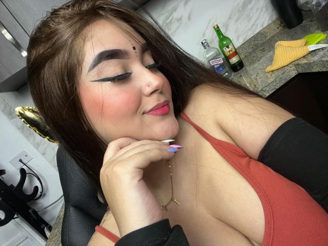 AnaShalimarr mit rosafarbenen Lippen, schwarzem Eyeliner und bunten, langen Nägeln (blau, lila, rosa, gelb), die ihre Hand küsst, braunes Haar, schwarzer Hintergrund, goldener Ohrring, kleiner Muttermal auf der Wange..