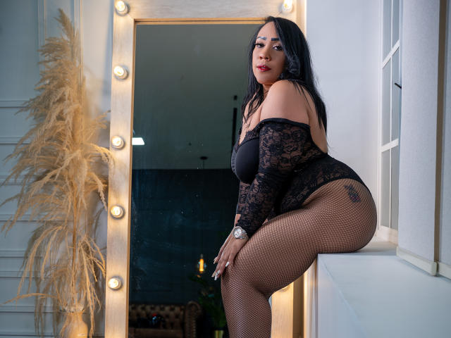 KarolWestt, com um body de renda preta, meias de rede e coxas grossas, apoiada em uma janela, exibindo seu grande bumbum, cabelo escuro e batom vermelho, usando um relógio de pulso..