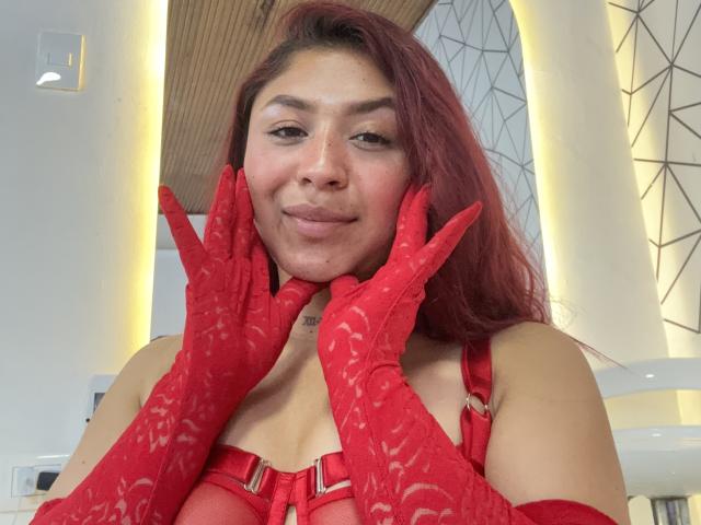 SophiaSaenz69 with red lace gloves and red lingerie, hands on her face, smiling slightly, red hair, light brown skin, medium tits, soft makeup, indoor setting, wood ceiling.