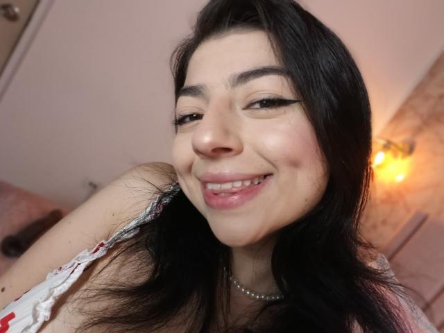 AnntonellaWatson with long black hair, black eyeliner, smiling, pink lips, pearl necklace, white dress with red pattern, and a cute face, looking at the camera.