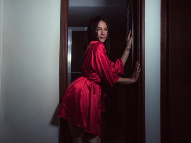 LizzeJoy with dark hair, wearing a shiny red satin dress, standing in a doorway, hands on frame, looking over shoulder, showing her ass, tattoo on right wrist.