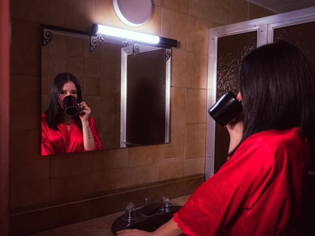 LizzeJoy met zwart haar, een rode satijnen blouse, die drinkt uit een zwarte beker, reflectie in de spiegel, onder een ronde lamp, badkamer, betegelde muren, wastafel met twee kranen, die de beker naar haar mond brengt..