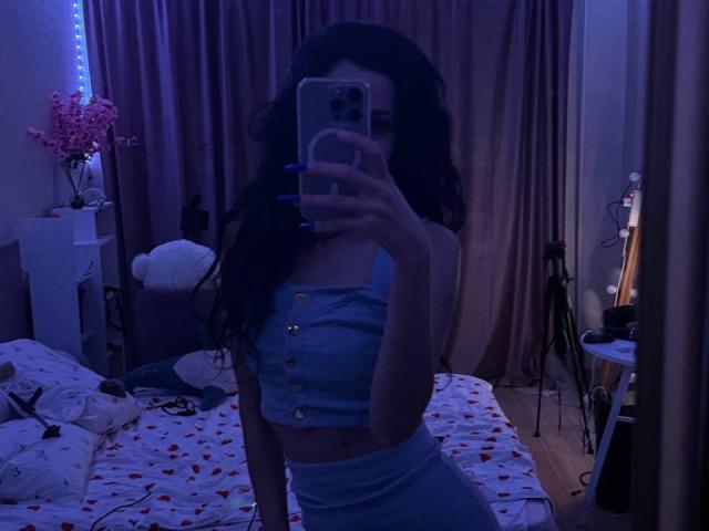 ShadowDance, avec ses cheveux noirs bouclés, son haut court bleu à boutons et son pantalon bleu taille haute, prend un selfie devant un miroir, montrant ses seins, avec des ongles bleus, dans une pièce éclairée en bleu sombre..