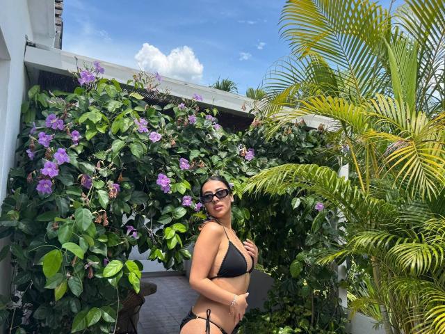Samantha Ross, con un bikini negro, mostrando sus senos, con gafas de sol, de pie frente a flores moradas y plantas verdes, hojas de palmera a la derecha, bajo un cielo azul..