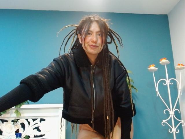 BeckyHuston with long dark dreadlocks, wearing a black zip-up jacket, bending forward, showing bare ass, smiling, blue wall, white decorative stand with candles in the background.